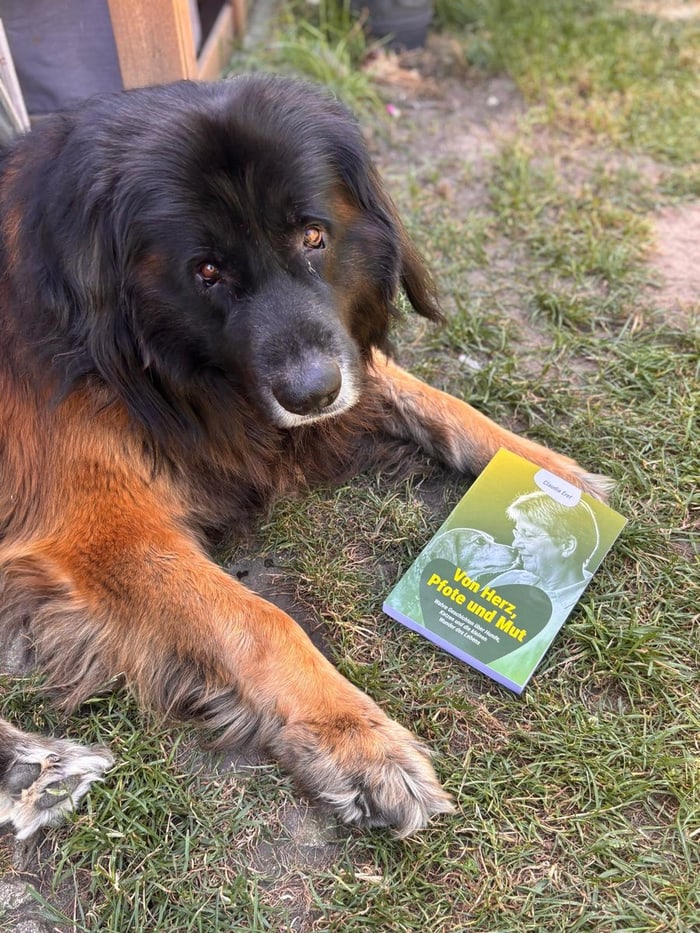 Der Hund Vaith liegt neben dem Buch „Von Herz, Pfote und Mut“ von Claudia Eret im Gras